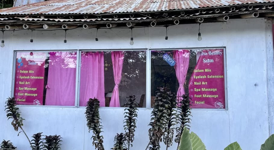 The white exterior of Nadiva Beauty salon in Waikabubak features three windows with bright pink curtains and an "open" sign, framed by plants and a rusty roof.