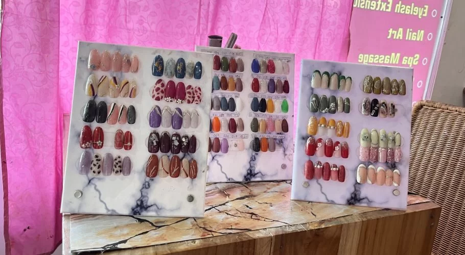 Three display boards showcase dozens of colorful artificial nails with intricate designs, patterns, and embellishments, set against a bright pink background.