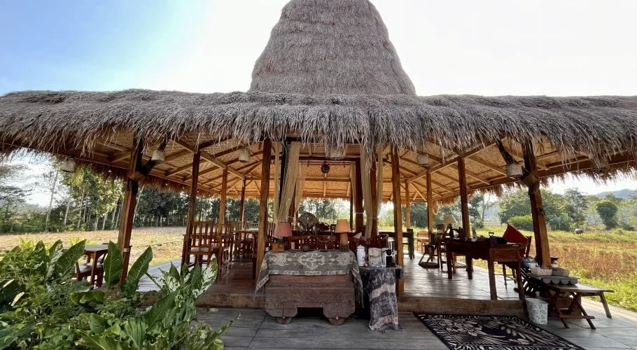 A large open-air restaurant pavilion with a prominent thatched roof and wooden structure overlooks green rice fields and distant trees under a bright sky.