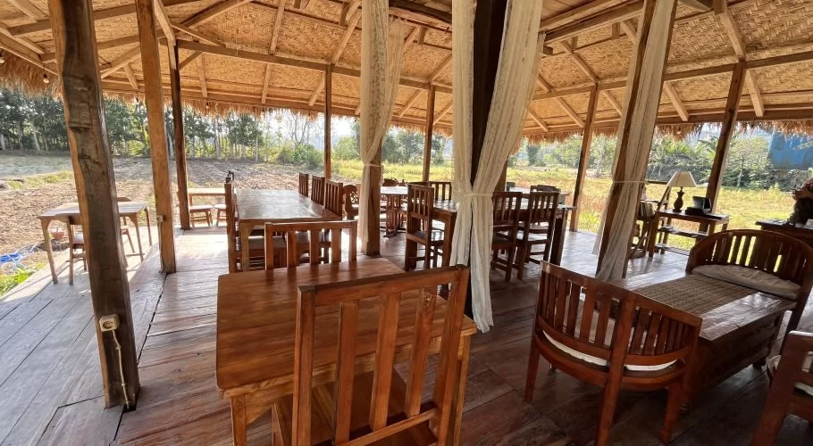 An open-air restaurant gazebo with a thatched roof and wooden tables and chairs overlooks vast, sunlit rice fields in West Sumba.