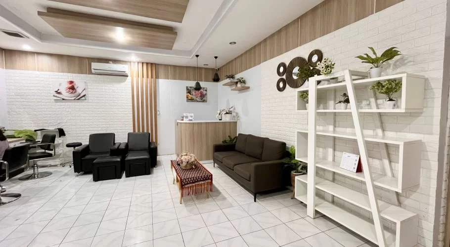 Interior of a modern spa reception area with white tiled floors, white brick walls, and wood accents, featuring a reception desk, black seating, and decorative plants.