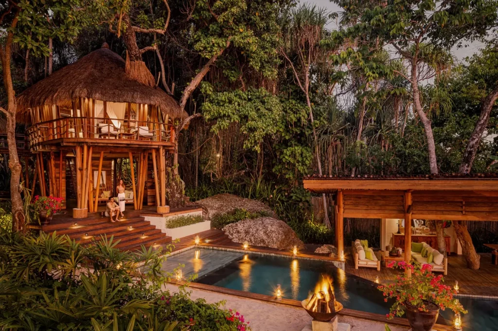 A luxurious thatched-roof treehouse villa with a couple on its steps, a private pool, and a lounge area, illuminated by warm lights at dusk amidst lush green jungle.