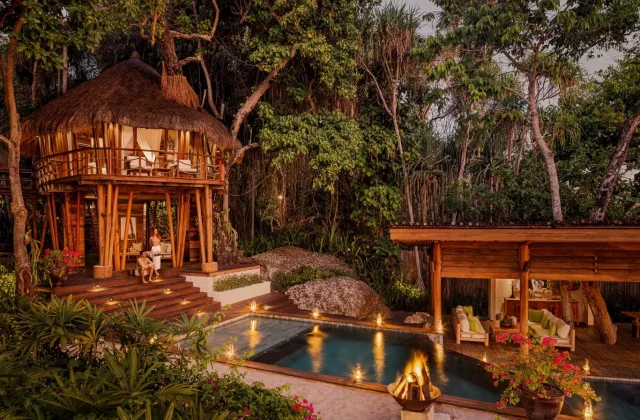 A luxurious thatched-roof treehouse villa with a couple on its steps, a private pool, and a lounge area, illuminated by warm lights at dusk amidst lush green jungle.