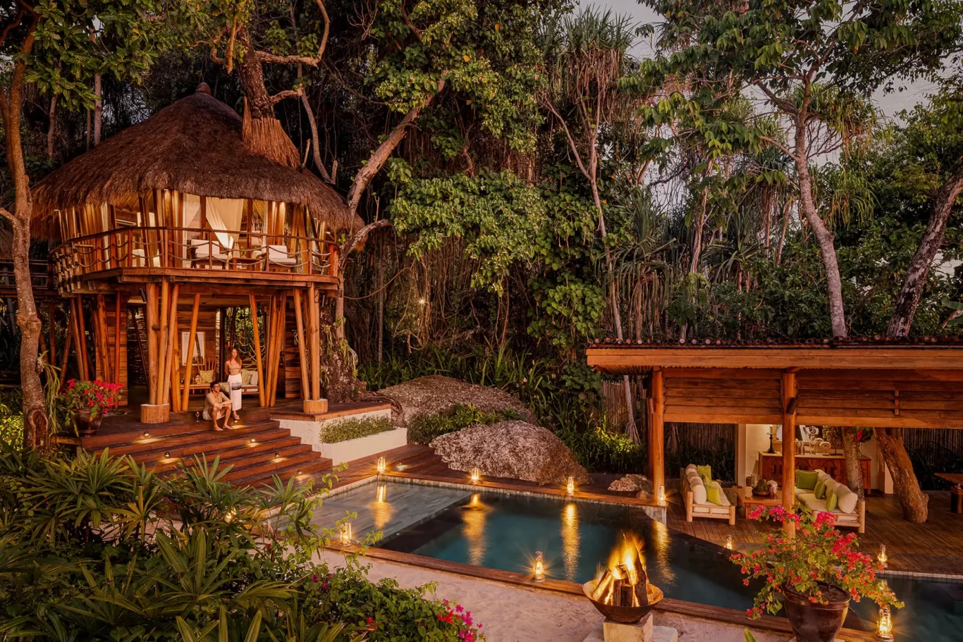 A luxurious thatched-roof treehouse villa with a couple on its steps, a private pool, and a lounge area, illuminated by warm lights at dusk amidst lush green jungle.