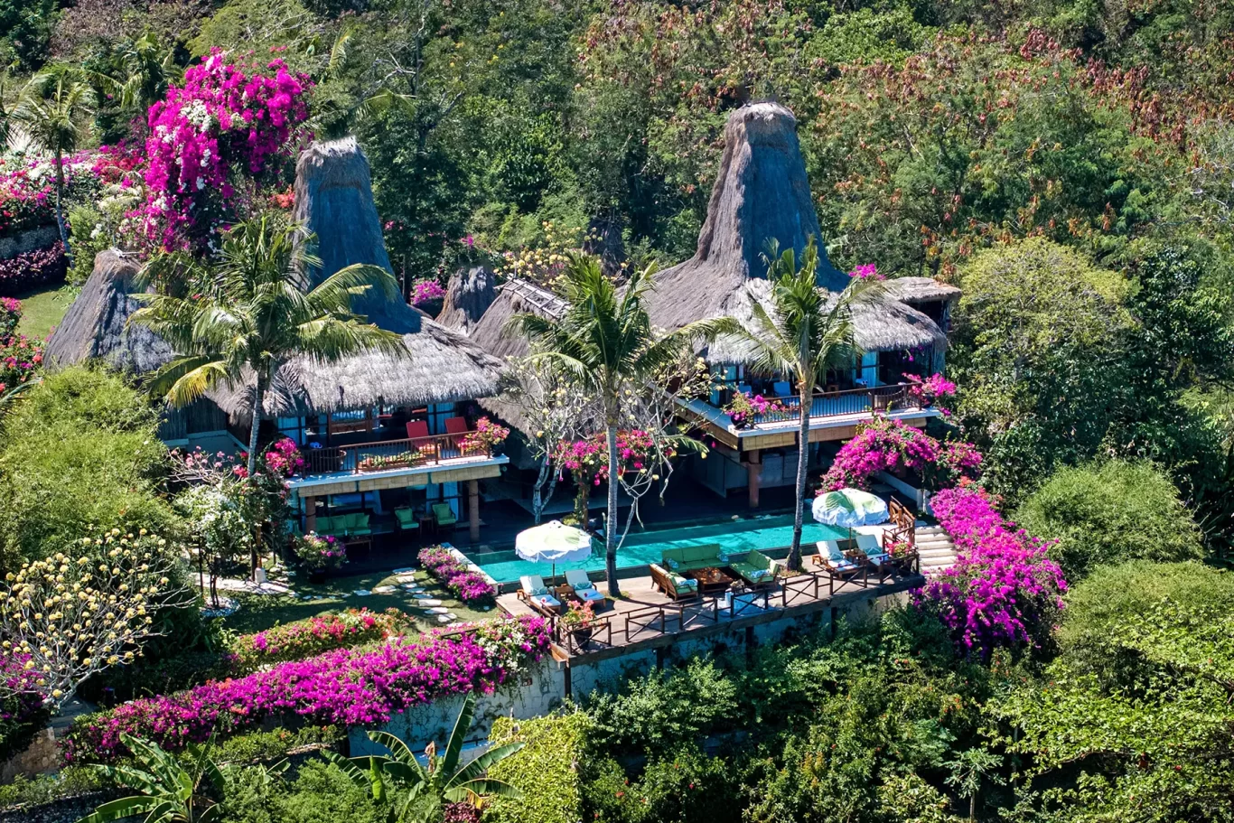 Aerial view of traditional thatched-roof villas at NIHI Sumba, surrounded by lush green jungle and vibrant pink bougainvillea, with a turquoise swimming pool.