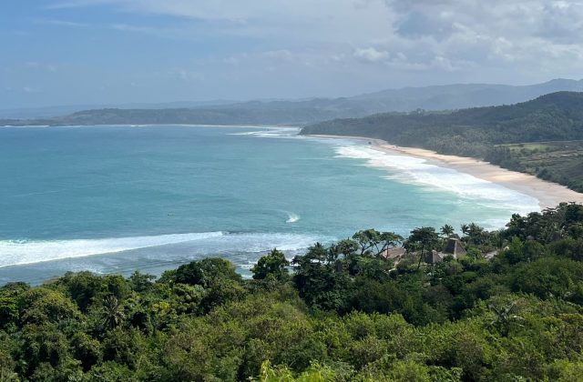 A panoramic view from Nihi Hill shows a long sandy beach with turquoise waves breaking, framed by lush green forests and distant hazy hills under a partly cloudy sky.