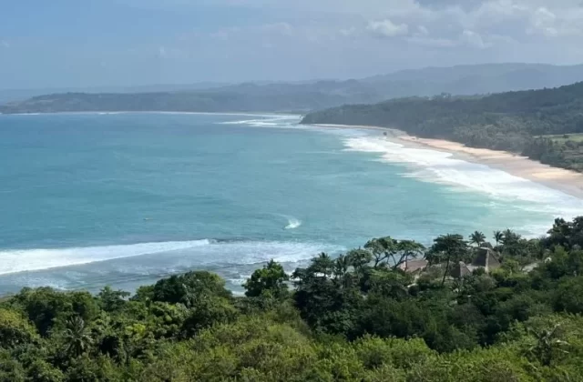 A panoramic view from Nihi Hill shows a long sandy beach with turquoise waves breaking, framed by lush green forests and distant hazy hills under a partly cloudy sky.