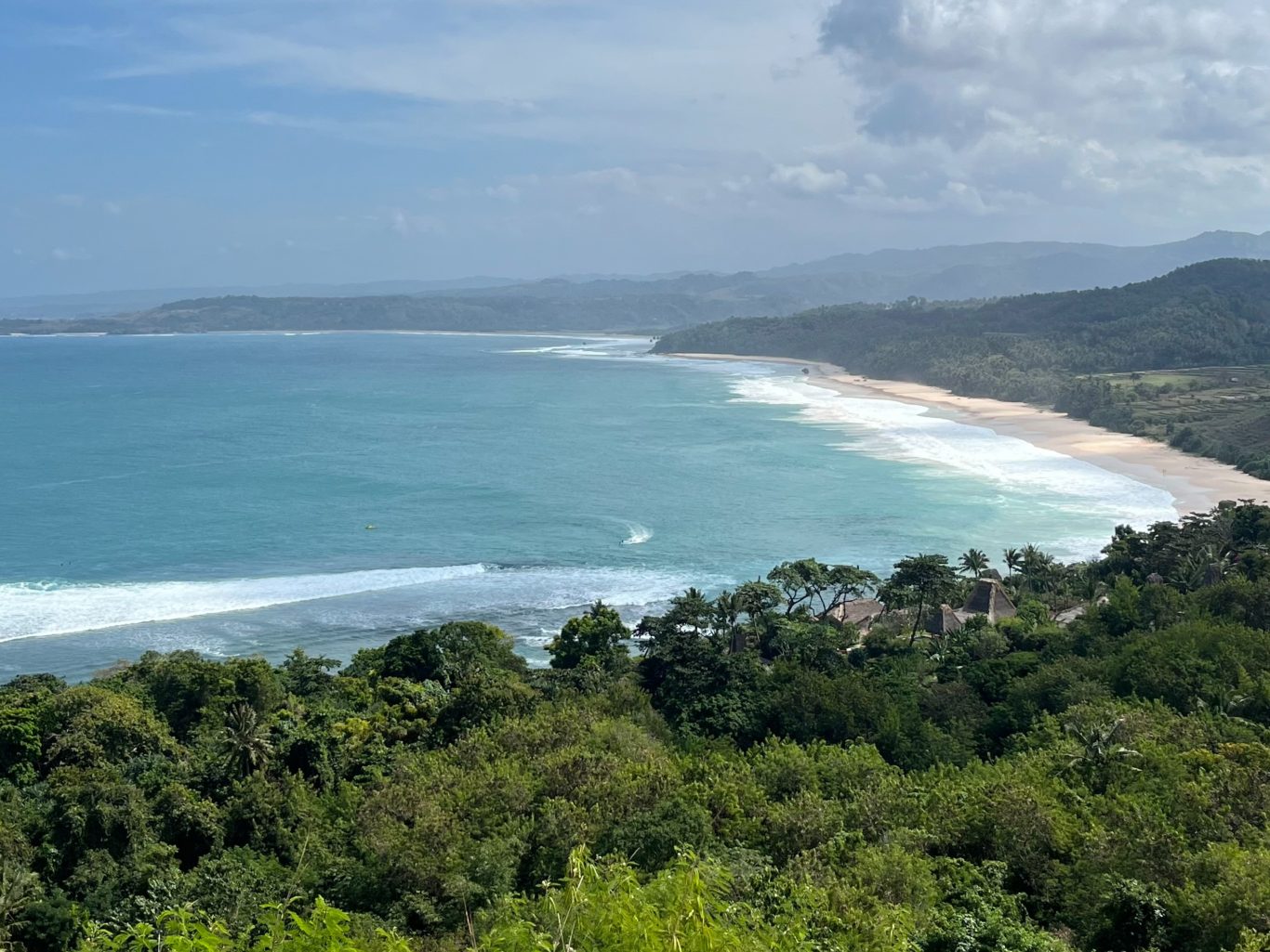 A panoramic view from Nihi Hill shows a long sandy beach with turquoise waves breaking, framed by lush green forests and distant hazy hills under a partly cloudy sky.