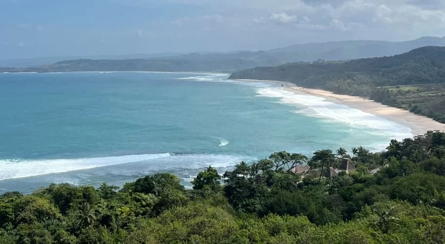 A panoramic view from Nihi Hill shows a long sandy beach with turquoise waves breaking, framed by lush green forests and distant hazy hills under a partly cloudy sky.