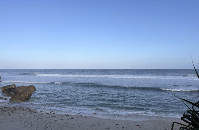 Ocean waves crash onto a sandy beach with large brown rocks on the left, under a clear blue sky, with dark foliage on the right.