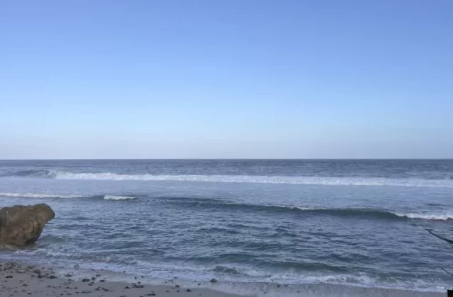 Ocean waves crash onto a sandy beach with large brown rocks on the left, under a clear blue sky, with dark foliage on the right.
