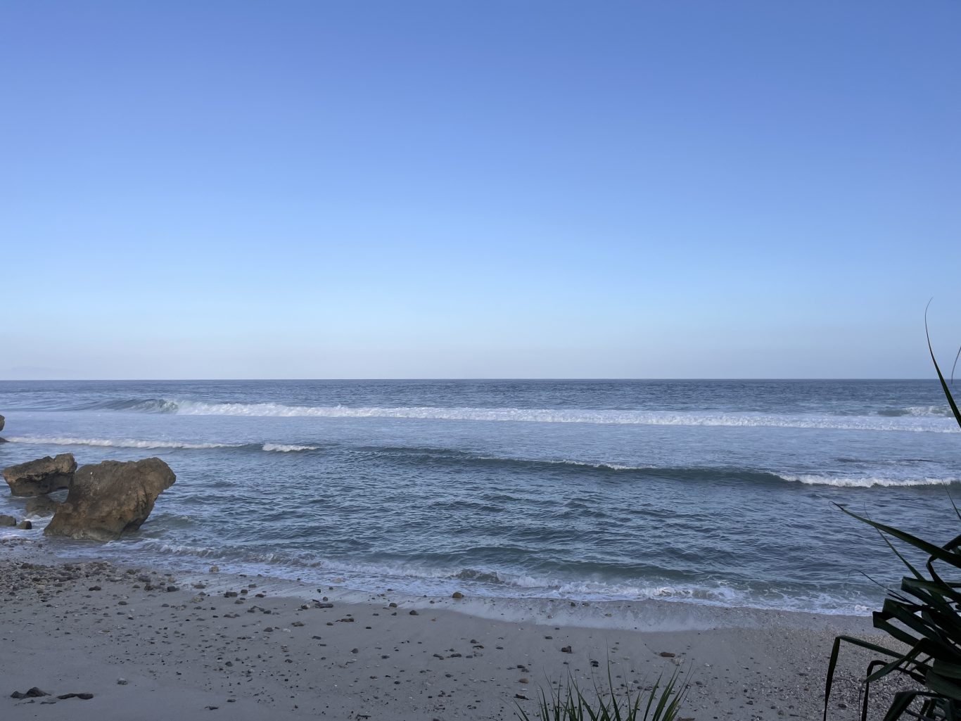 Ocean waves crash onto a sandy beach with large brown rocks on the left, under a clear blue sky, with dark foliage on the right.