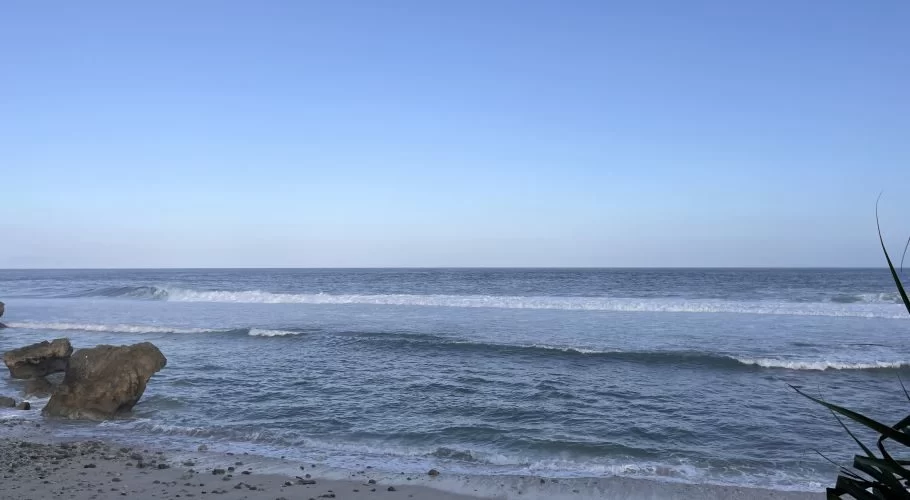 Ocean waves crash onto a sandy beach with large brown rocks on the left, under a clear blue sky, with dark foliage on the right.