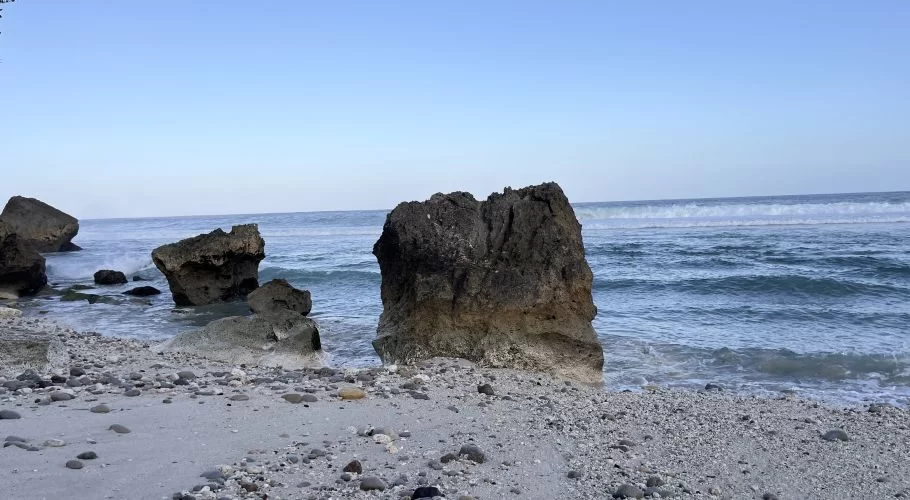 Large dark rocks stand on a light sandy beach with scattered pebbles, as blue ocean waves gently break under a clear sky.