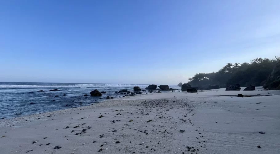 Nihi Oka Beach features white sand with dark pebbles, blue ocean waves breaking around large rocks, and a lush green treeline under a clear blue sky.
