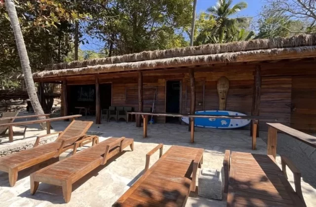 A rustic wooden building with a thatched roof, a blue surfboard, and wooden lounge chairs are on a paved area, tropical trees visible.