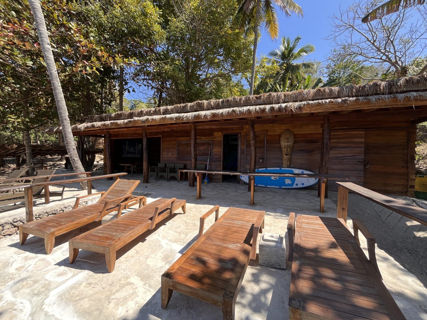 A rustic wooden building with a thatched roof, a blue surfboard, and wooden lounge chairs are on a paved area, tropical trees visible.