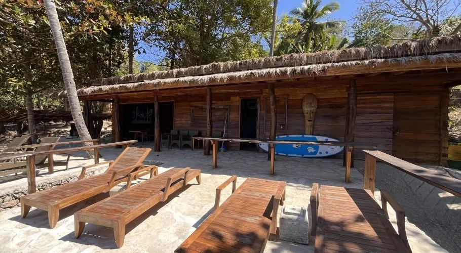 A rustic wooden building with a thatched roof, a blue surfboard, and wooden lounge chairs are on a paved area, tropical trees visible.