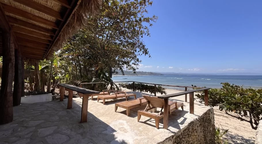 A sunny outdoor sunbathing area with wooden lounge chairs on a stone patio overlooks a sandy beach and the blue ocean at Rua Beach, Sumba.