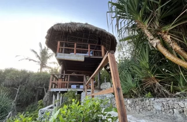 A two-story, open-air spa structure with a thatched roof stands among lush green tropical foliage under a bright sky.