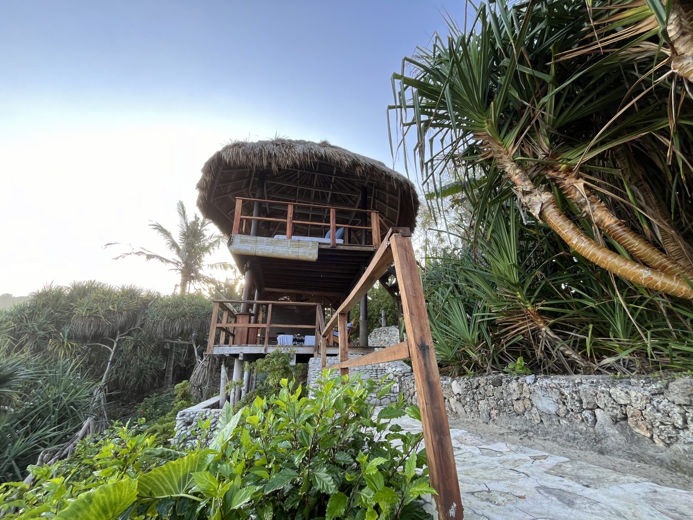 A two-story, open-air spa structure with a thatched roof stands among lush green tropical foliage under a bright sky.
