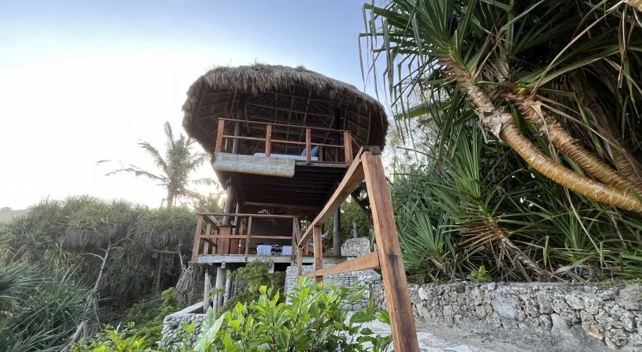 A two-story, open-air spa structure with a thatched roof stands among lush green tropical foliage under a bright sky.