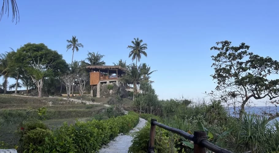 A stone path winds through lush green tropical foliage towards a two-story, thatched-roof spa building on a hill under a clear blue sky, with the ocean visible.