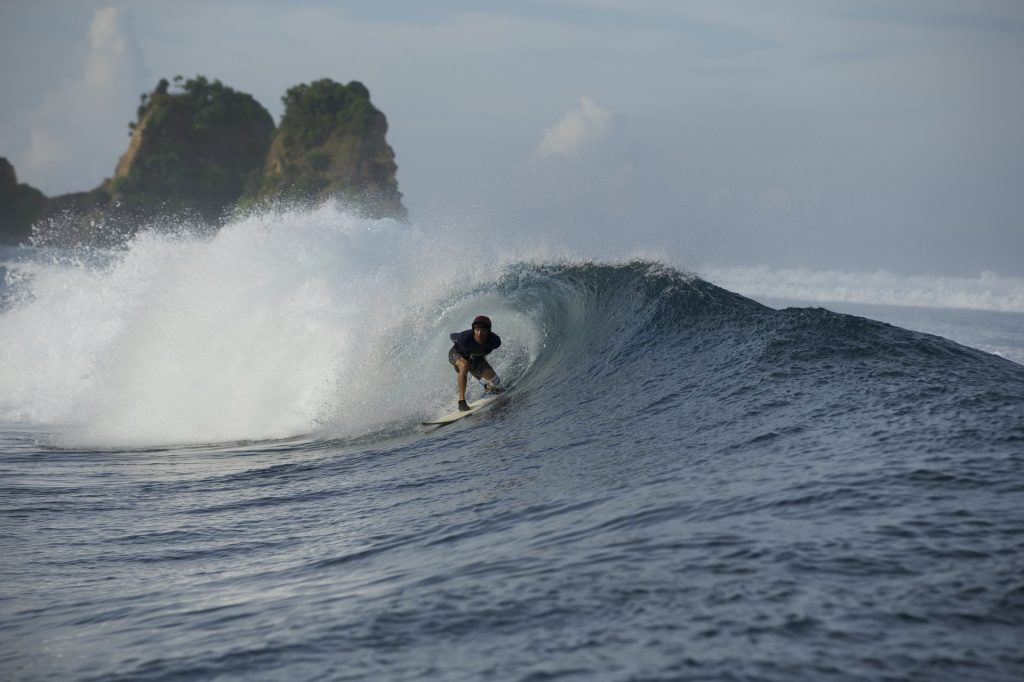 A surfer in dark attire rides inside a powerful, breaking barrel wave on dark blue ocean waters, with green rocky islands in the background under a cloudy sky.
