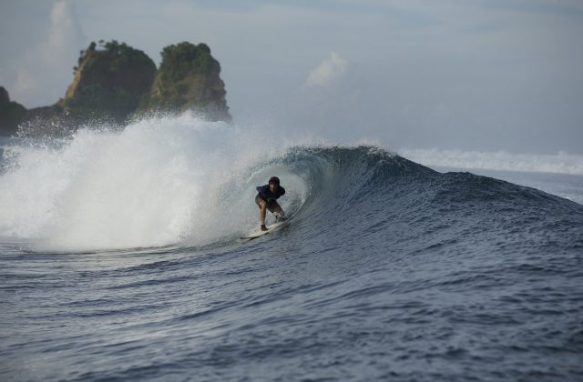 A surfer in dark attire rides inside a powerful, breaking barrel wave on dark blue ocean waters, with green rocky islands in the background under a cloudy sky.