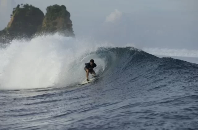 A surfer in dark attire rides inside a powerful, breaking barrel wave on dark blue ocean waters, with green rocky islands in the background under a cloudy sky.
