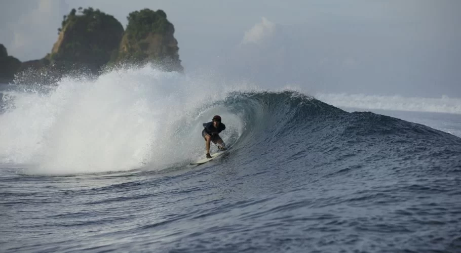 A surfer in dark attire rides inside a powerful, breaking barrel wave on dark blue ocean waters, with green rocky islands in the background under a cloudy sky.