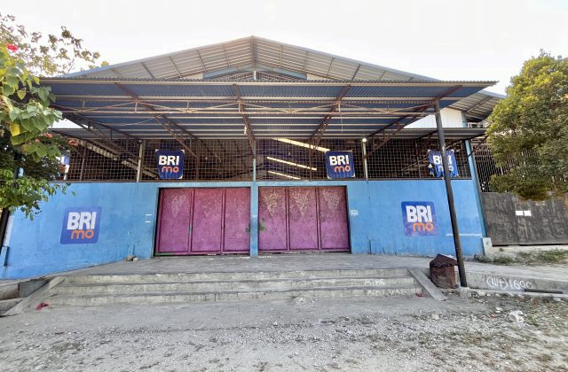 The exterior of Oasis Delta Futsal in Waikabubak features a blue wall with faded pink doors, a corrugated metal roof, and BRImo logos, with concrete steps leading up.
