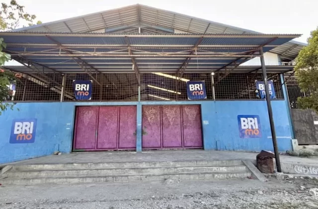 The exterior of Oasis Delta Futsal in Waikabubak features a blue wall with faded pink doors, a corrugated metal roof, and BRImo logos, with concrete steps leading up.