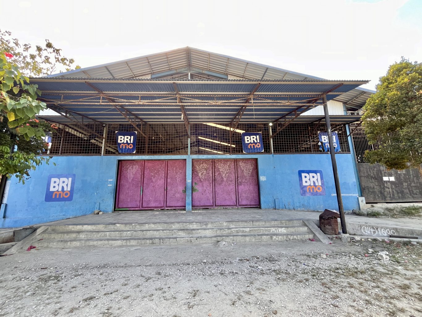 The exterior of Oasis Delta Futsal in Waikabubak features a blue wall with faded pink doors, a corrugated metal roof, and BRImo logos, with concrete steps leading up.