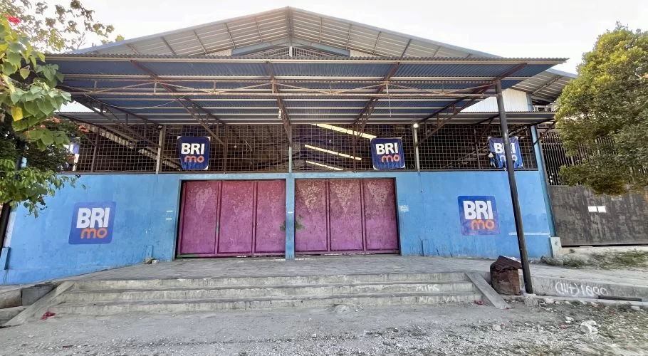 The exterior of Oasis Delta Futsal in Waikabubak features a blue wall with faded pink doors, a corrugated metal roof, and BRImo logos, with concrete steps leading up.