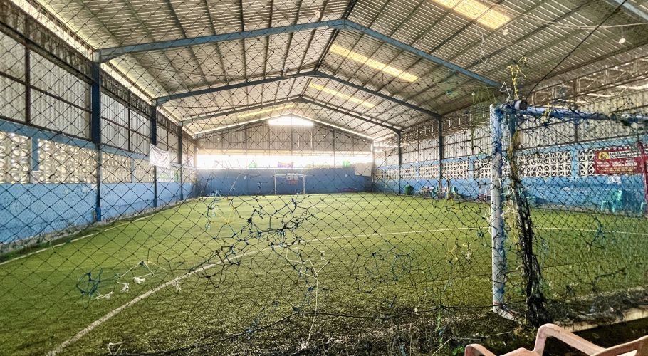 An empty indoor futsal court with green artificial turf, blue walls, a metal roof, and a goal at the far end, viewed through a foreground net.