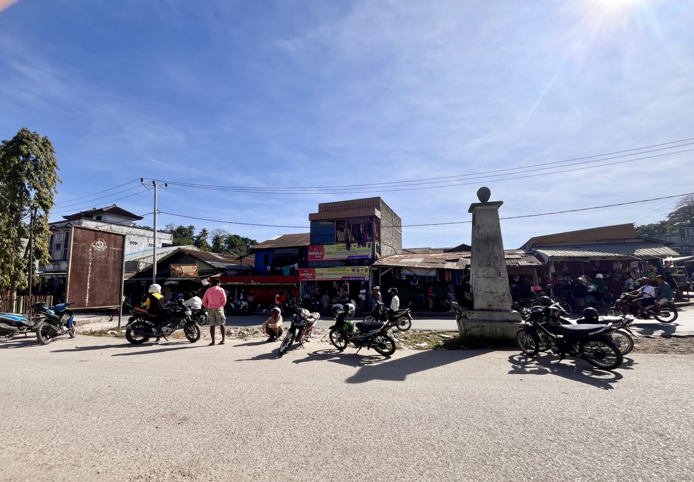 Motorcycle taxis and people gather on a sunny street in Waikabubak, Sumba, with various buildings and a stone monument in the background.