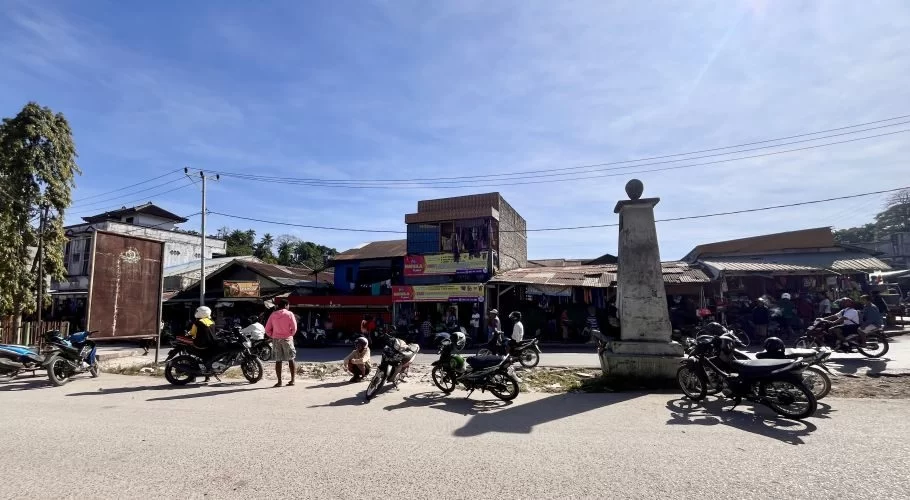 Motorcycle taxis and people gather on a sunny street in Waikabubak, Sumba, with various buildings and a stone monument in the background.