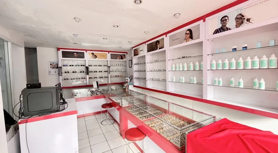 The interior of Optik International in Waikabubak features white walls, red and white display cases filled with eyeglasses frames, and shelves stocked with contact lens solutions.