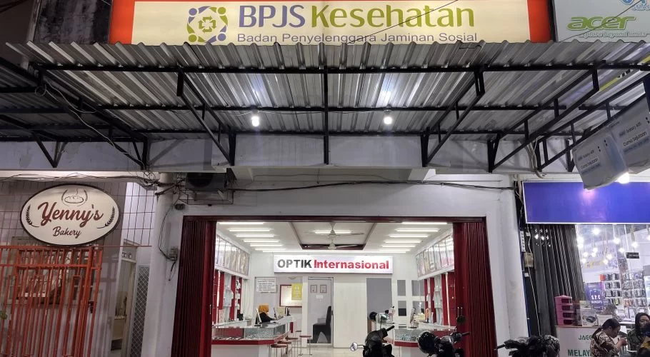 The brightly lit storefront of Optik Internasional in Waingapu, Sumba, is visible with a BPJS Kesehatan sign above and Yenny's Bakery sign to the left.