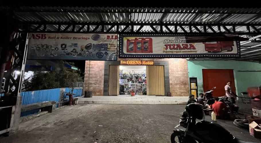 A brightly lit motorcycle repair shop, Orens Motor, is seen at night with mechanics working on bikes and advertising banners above the entrance.