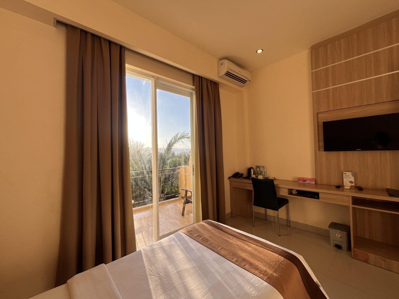 A modern guest room at Padadita Beach Hotel features a bed, desk, wall-mounted TV, and sliding glass doors to a balcony overlooking palm trees under a bright sky.