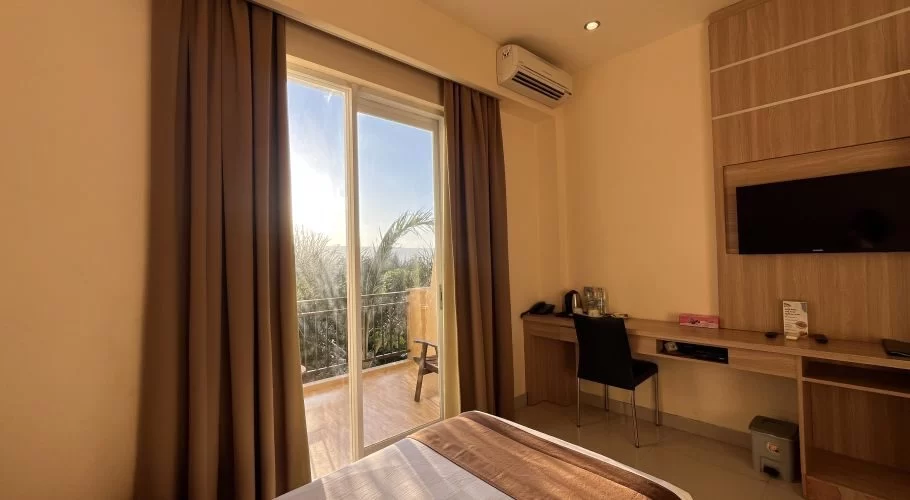 A modern guest room at Padadita Beach Hotel features a bed, desk, wall-mounted TV, and sliding glass doors to a balcony overlooking palm trees under a bright sky.