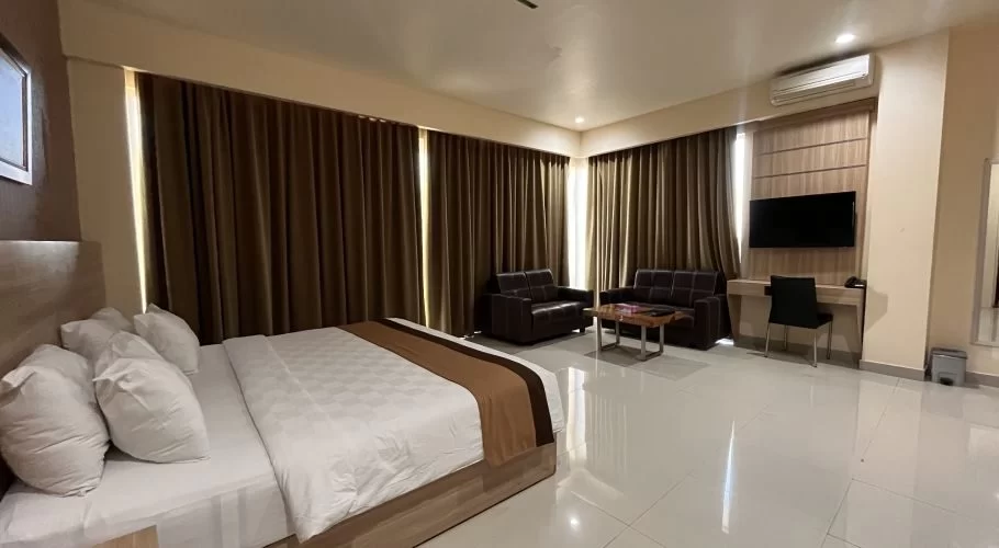 A hotel room with a large bed, two brown sofas, a coffee table, and a wall-mounted TV. Brown curtains line the walls.