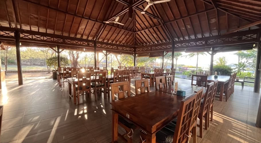 Spacious open-air dining area at Padadita Beach Hotel with dark wooden tables, chairs, and a high ceiling. Sunlight casts shadows on the tiled floor, with trees and the ocean visible outside.