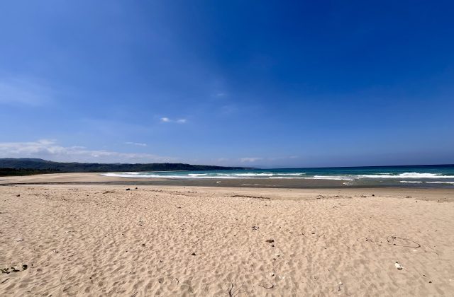 A vast, light tan sandy beach stretches towards the turquoise ocean with gentle waves under a bright blue sky, backed by dark green hills.