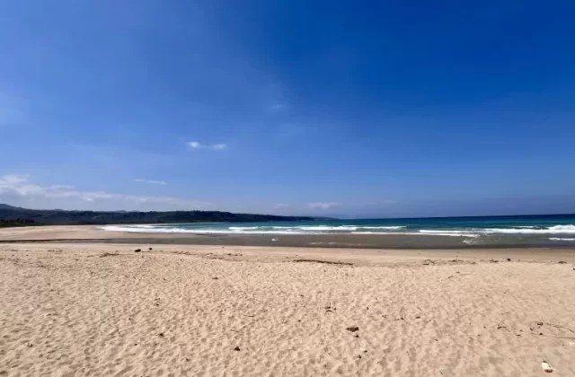 A vast, light tan sandy beach stretches towards the turquoise ocean with gentle waves under a bright blue sky, backed by dark green hills.