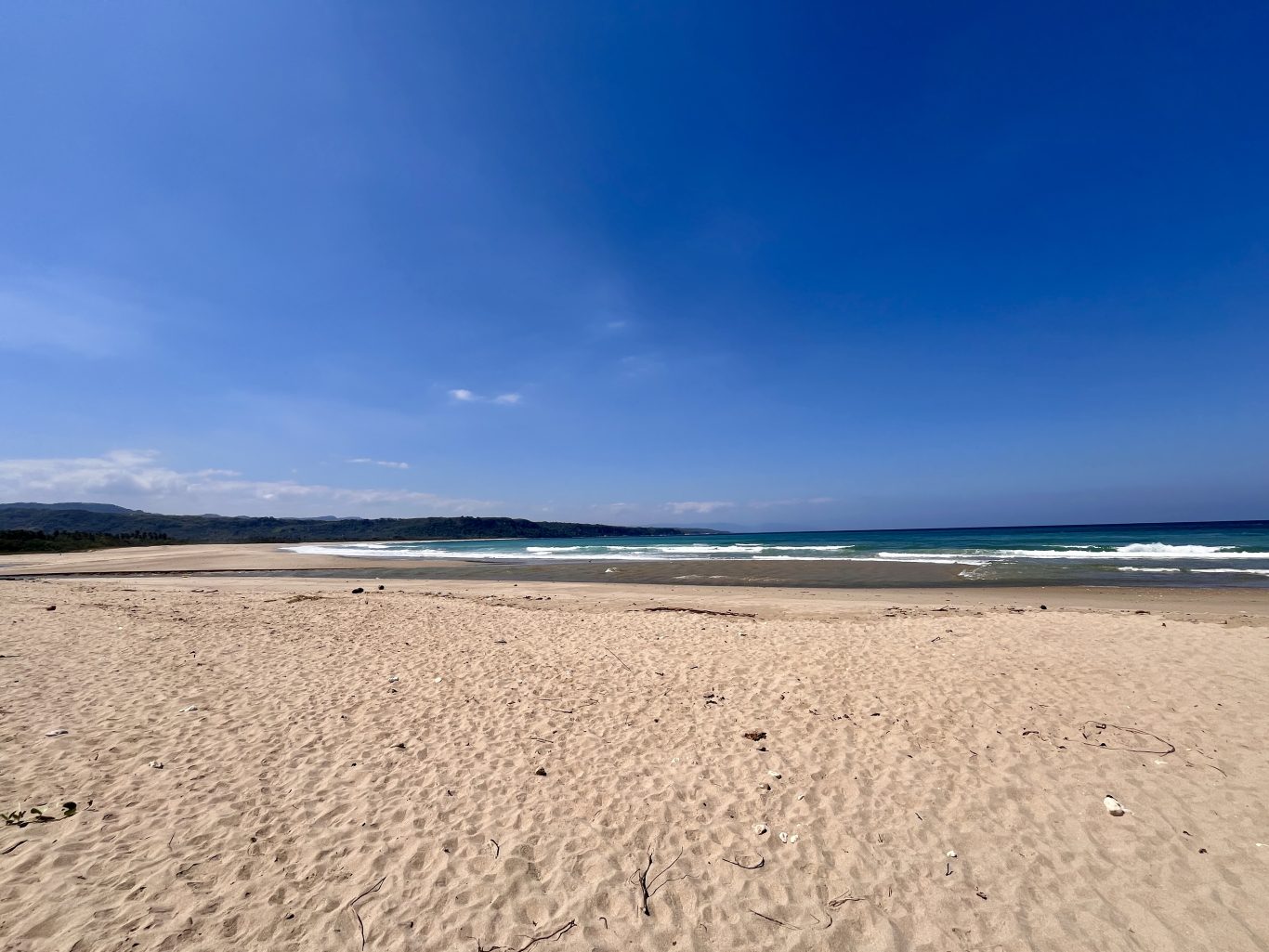 A vast, light tan sandy beach stretches towards the turquoise ocean with gentle waves under a bright blue sky, backed by dark green hills.