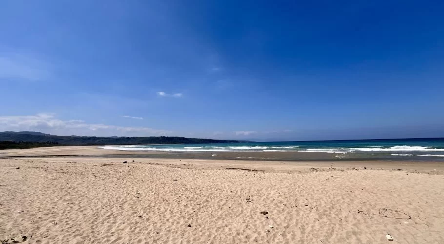 A vast, light tan sandy beach stretches towards the turquoise ocean with gentle waves under a bright blue sky, backed by dark green hills.