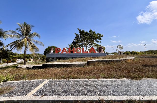 Large red letters spelling "PAHOLA" stand on a concrete base amidst dry grass, palm trees, and other greenery under a clear blue sky.